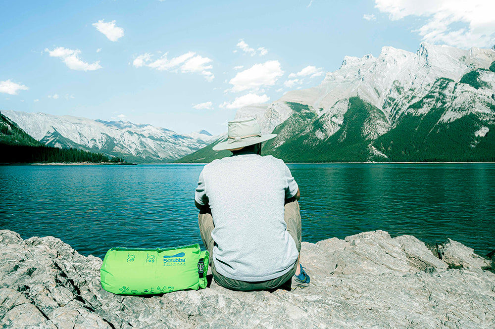 Man sitting on rocks on edge of lake with a Scrubba green wash bag